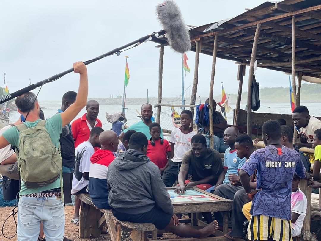 Sound recordist with a boom microphone capturing community life at a fishing shelter in Dixcove