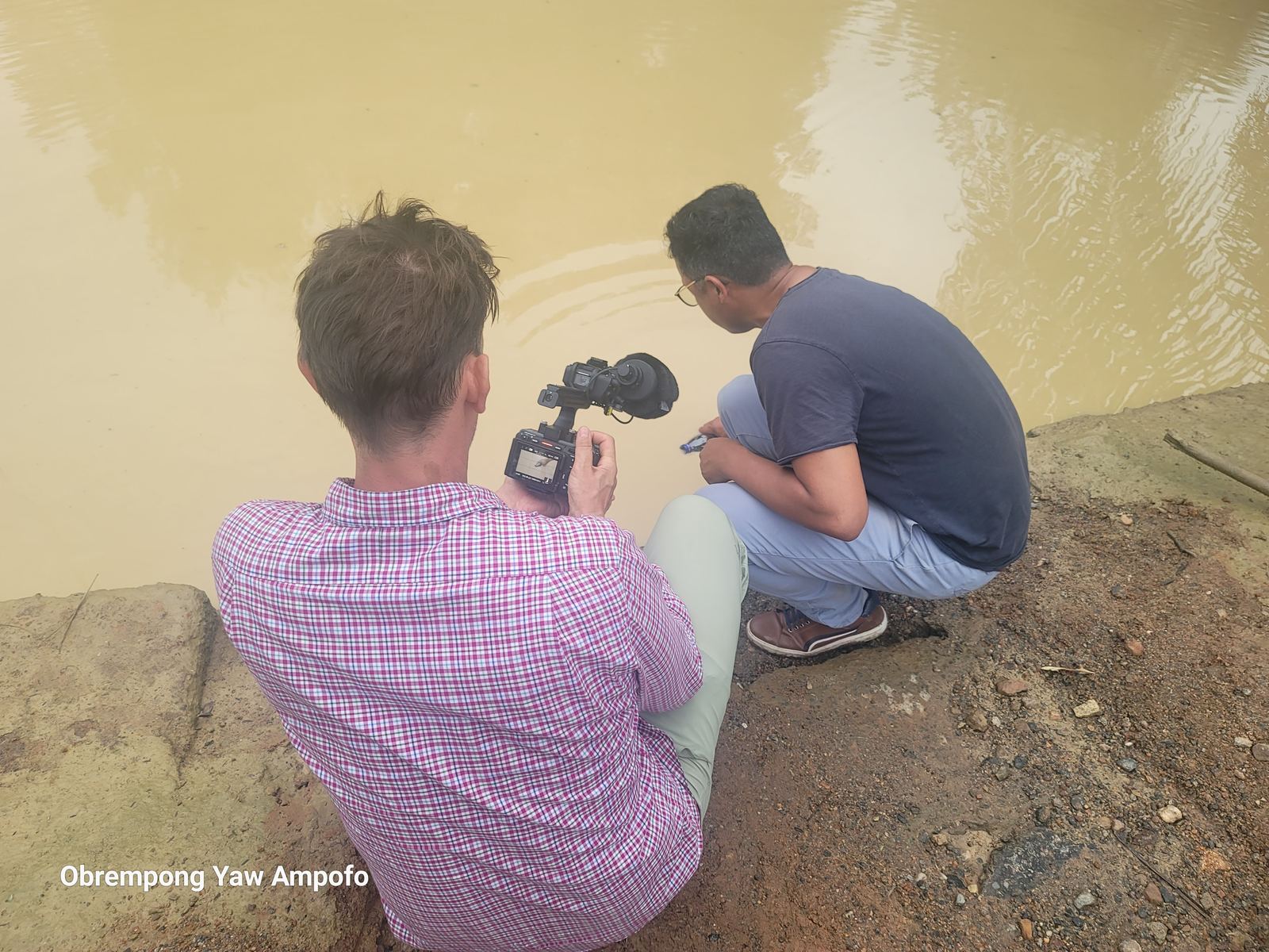 Two men crouched at a riverbank collecting a water sample from the Whin River